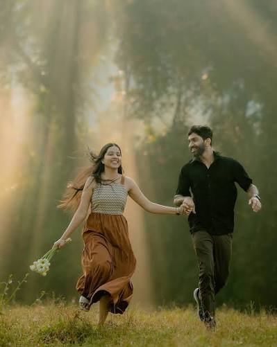 A smiling couple holding hands and running through a sunlit meadow surrounded by trees.